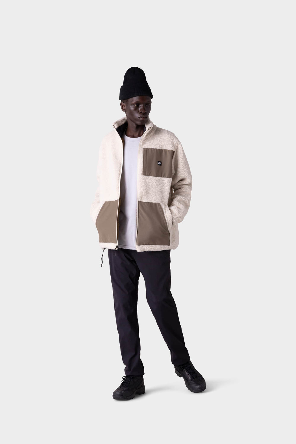 A man wearing the 686 Men's Everywhere® Merino-Lined Pant - Slim Fit stands against a plain grey background, styled for cold weather with a black beanie, cream and beige fleece jacket, white t-shirt, and black shoes.