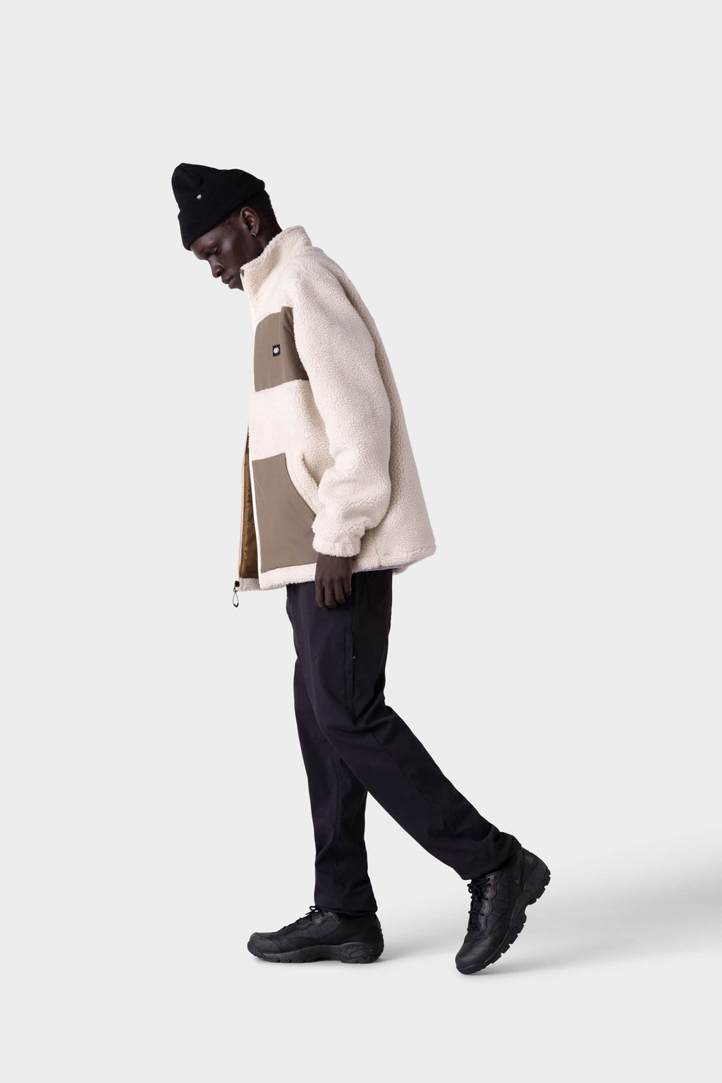 A person in a warm cream and brown fleece jacket, black beanie, and black shoes stands in profile against a plain light background, wearing 686 Men's Everywhere® Merino-Lined Pant - Slim Fit.