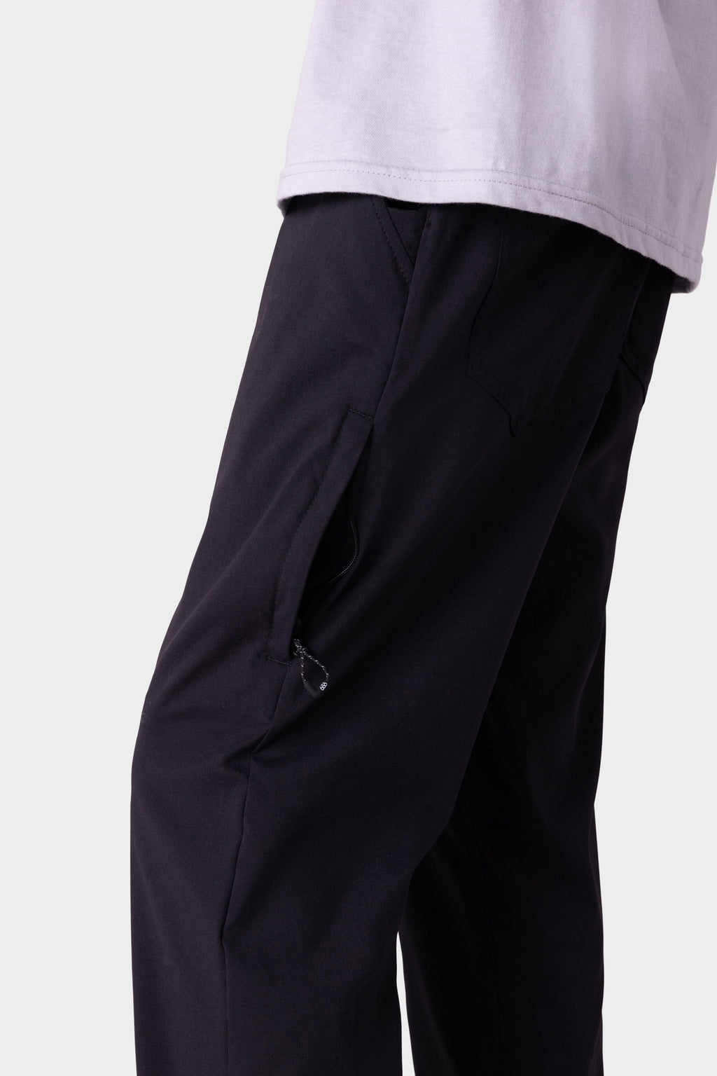 A close-up of a person wearing the 686 Men's Everywhere® Merino-Lined Pant - Slim Fit in black, featuring a zippered pocket for warmth, paired with a light shirt. The image shows the lower torso and upper legs against a plain background.
