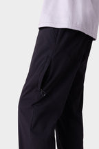 A close-up of a person wearing the 686 Men's Everywhere® Merino-Lined Pant - Slim Fit in black, featuring a zippered pocket for warmth, paired with a light shirt. The image shows the lower torso and upper legs against a plain background.