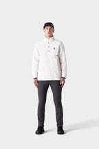 A man faces forward against a plain light gray background, dressed for colder conditions in a black cap, white long-sleeve buttoned pullover, black shoes, and 686 Men's Everywhere® Merino-Lined Pant - Slim Fit in dark gray.