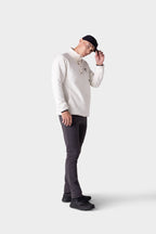 A person stands against a plain light background, wearing the 686 Men's Everywhere® Merino-Lined Pant - Slim Fit by 686 along with a white long-sleeve shirt, black shoes, and a dark cap, slightly tilting their head while holding the brim of their cap.