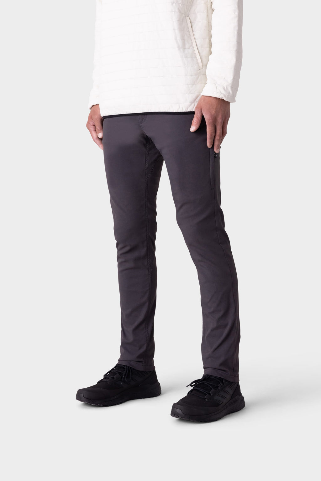 Wearing 686 Men's Everywhere® Merino-Lined Pant - Slim Fit in dark gray, paired with a white long-sleeve and black sneakers, a person stands against a plain light gray background. The image is cropped above the shoulders.