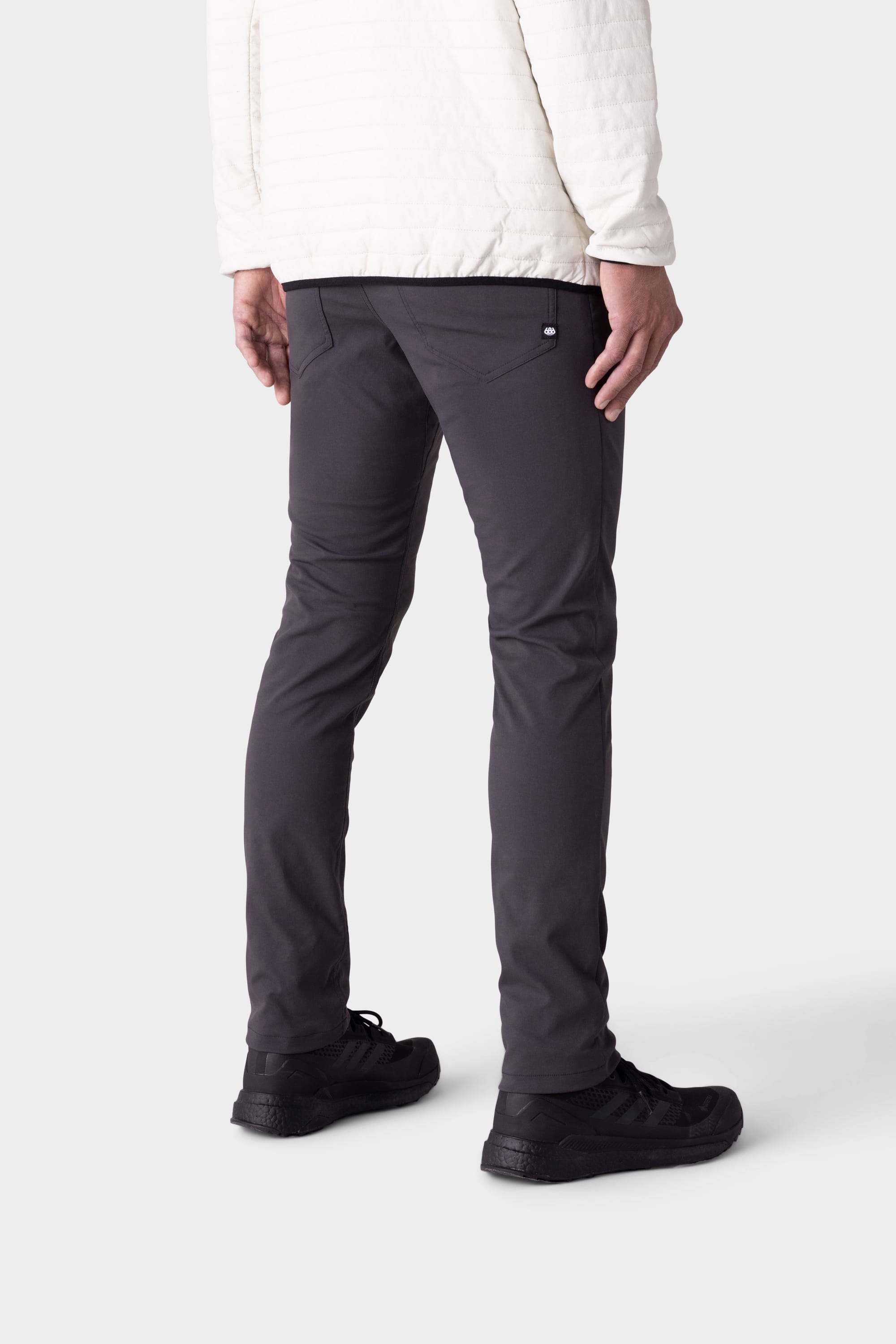 A person stands facing away in 686 Men's Everywhere® Merino-Lined Pant - Slim Fit (dark gray), pairing warmth and style, with a white quilted jacket and black sneakers, shown from the back and side against a plain light background.