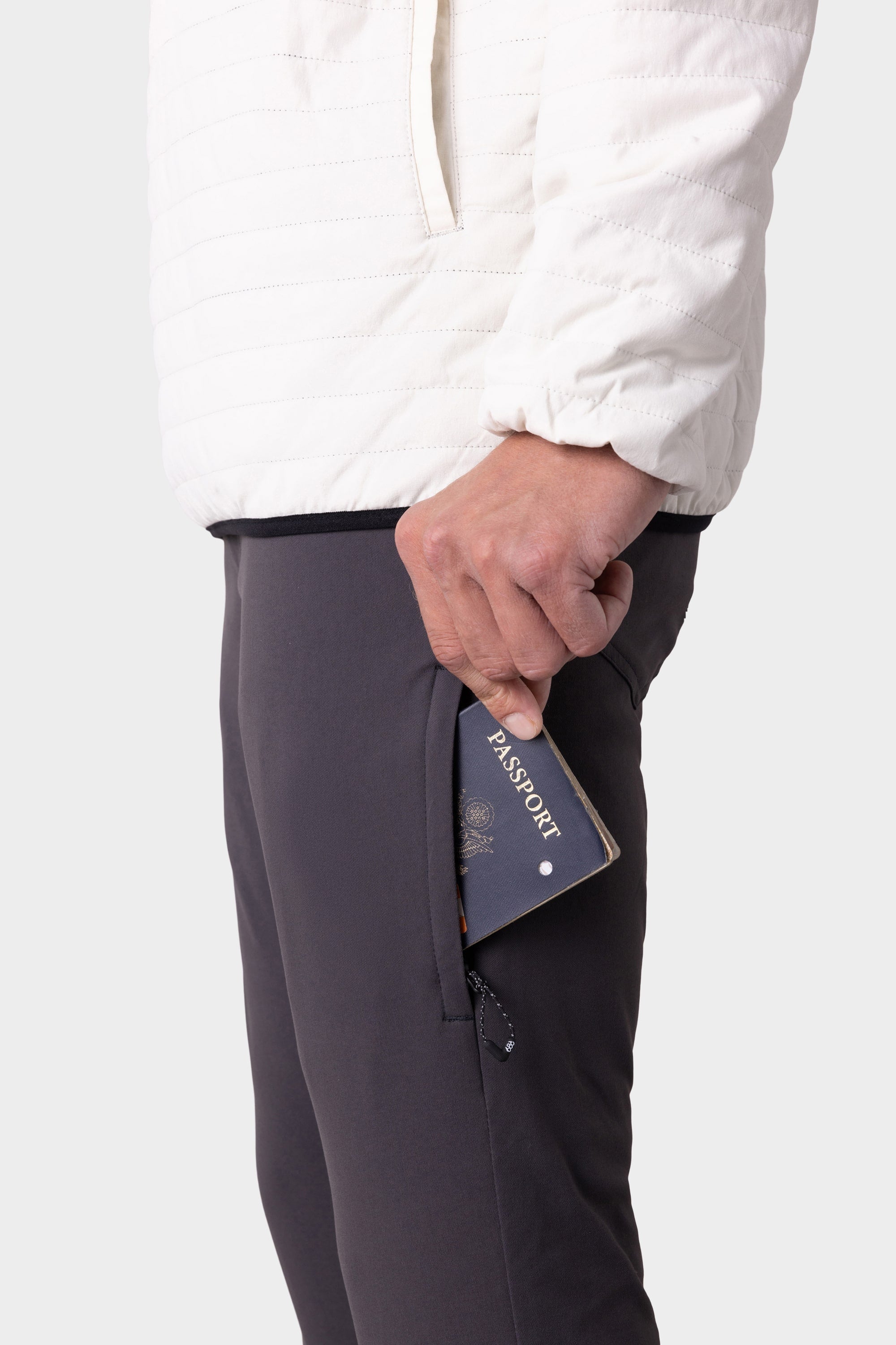 Wearing the 686 Men's Everywhere® Merino-Lined Pant - Slim Fit, a person dressed for cold weather puts a dark blue passport into the zippered pocket of their pants.