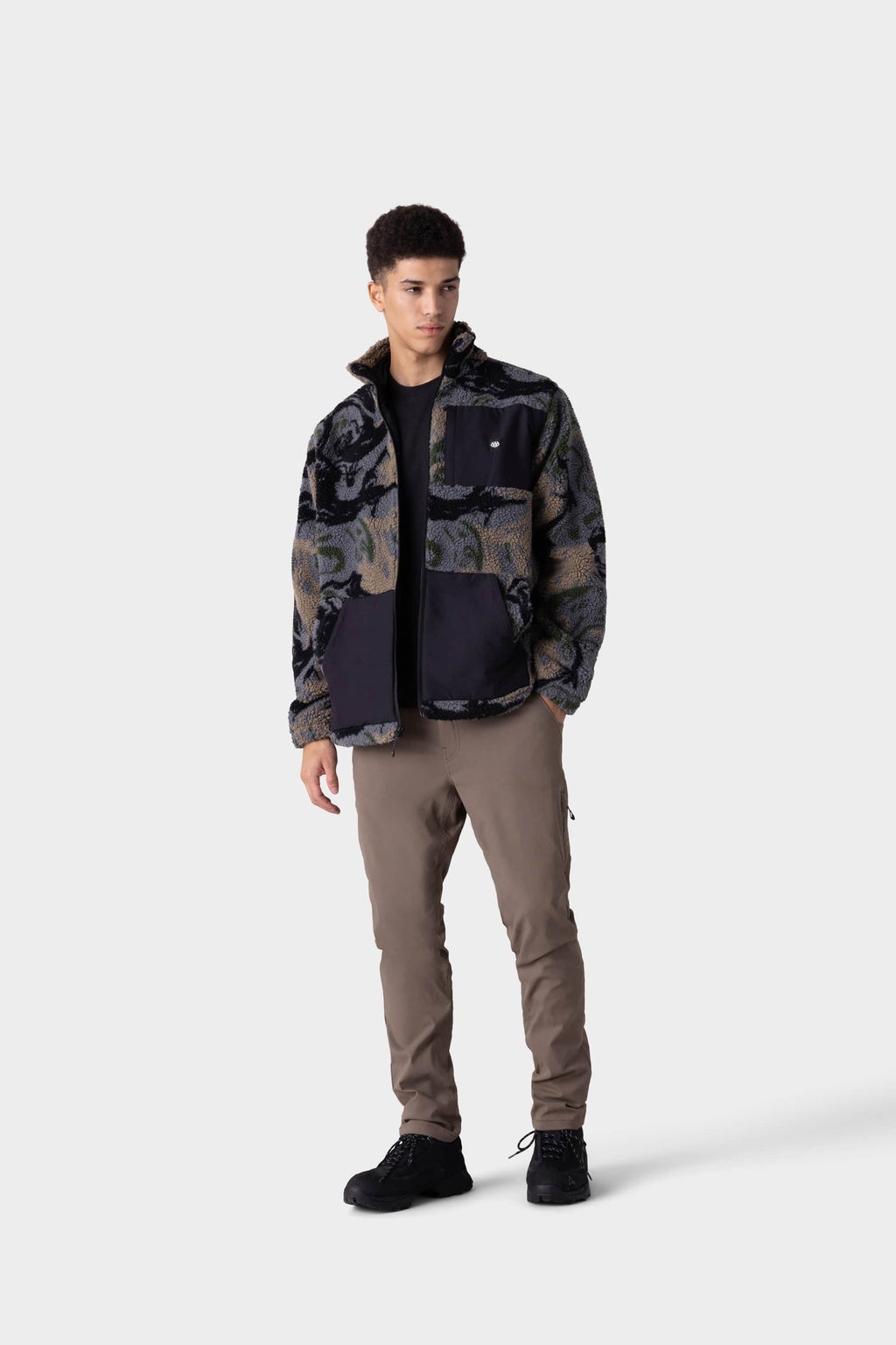 A person stands facing forward against a plain light background, wearing khaki 686 Men's Everywhere® Merino-Lined Pant (Slim Fit), a patterned fleece jacket, black shirt, and black shoes to stay warm in colder conditions.