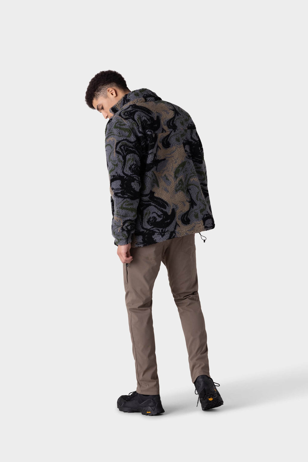 A man faces away, head down, wearing a warm patterned fleece jacket, 686 Men's Everywhere® Merino-Lined Pant - Slim Fit in light brown by 686, and black shoes against a plain light gray background.