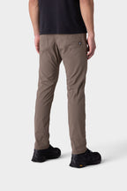 A person dressed for colder weather in 686 Men's Everywhere® Merino-Lined Pant - Slim Fit (taupe), a black shirt, and black shoes stands facing away against a plain light gray background.