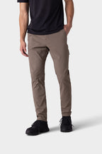 A person in 686 Men's Everywhere® Merino-Lined Pant - Slim Fit (taupe), a black t-shirt, and black shoes—ideal for dry, cold weather—stands with one hand in their pocket against a plain light gray background. Head is not visible.