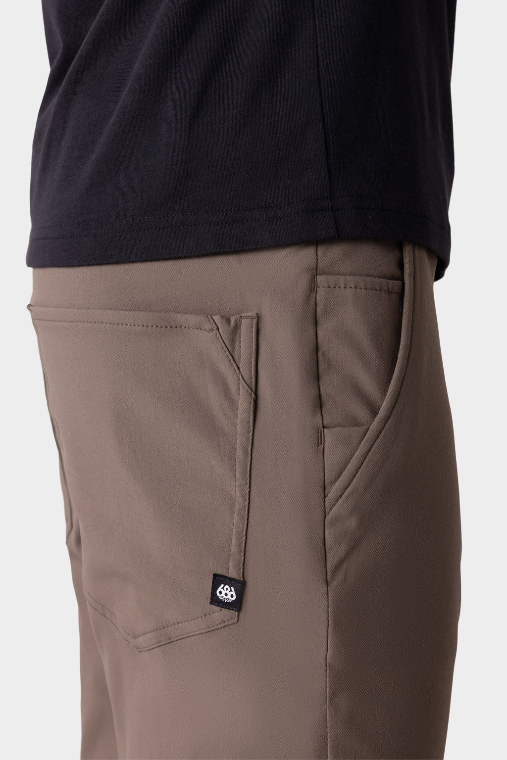 A close-up side view shows a person wearing 686 Men's Everywhere® Merino-Lined Pant - Slim Fit in taupe, featuring a front pocket and black label—perfect for warmth. Paired with a dark shirt, they're set against a plain, light background.
