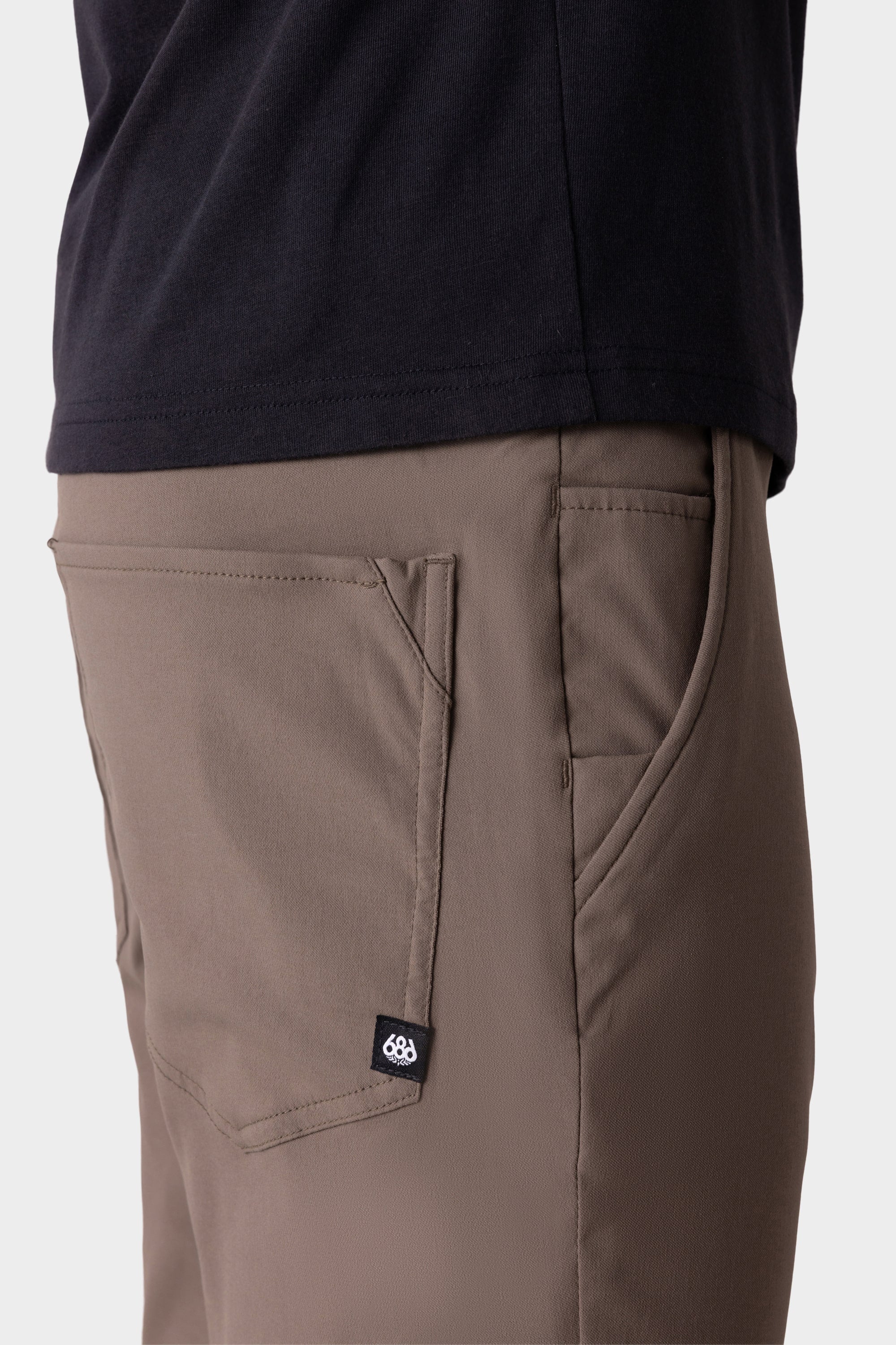 A close-up side view shows a person wearing 686 Men's Everywhere® Merino-Lined Pant - Slim Fit in taupe, featuring a front pocket and black label—perfect for warmth. Paired with a dark shirt, they're set against a plain, light background.