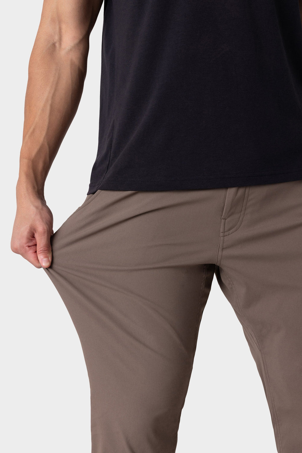 A person in 686 Men's Everywhere® Merino-Lined Pant - Slim Fit (brown) demonstrates the flexible fabric with one hand. The warm pant is visible from waist to knees; the person pairs it with a black shirt.