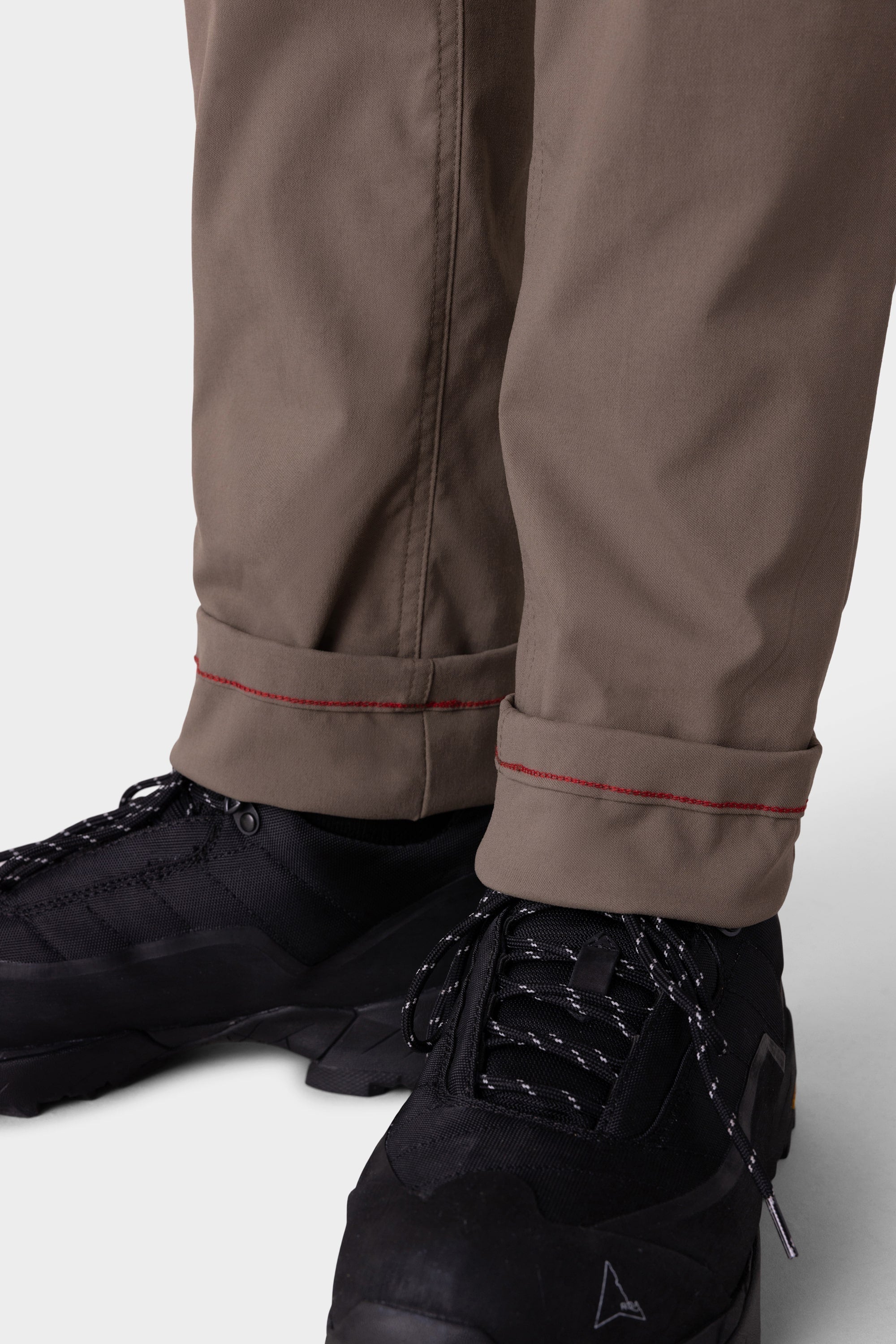 Close-up of a person in 686 Men's Everywhere® Merino-Lined Pant - Slim Fit in light brown with red-stitched cuffs, paired with black athletic shoes—designed by 686 to keep you warm and dry in colder conditions.