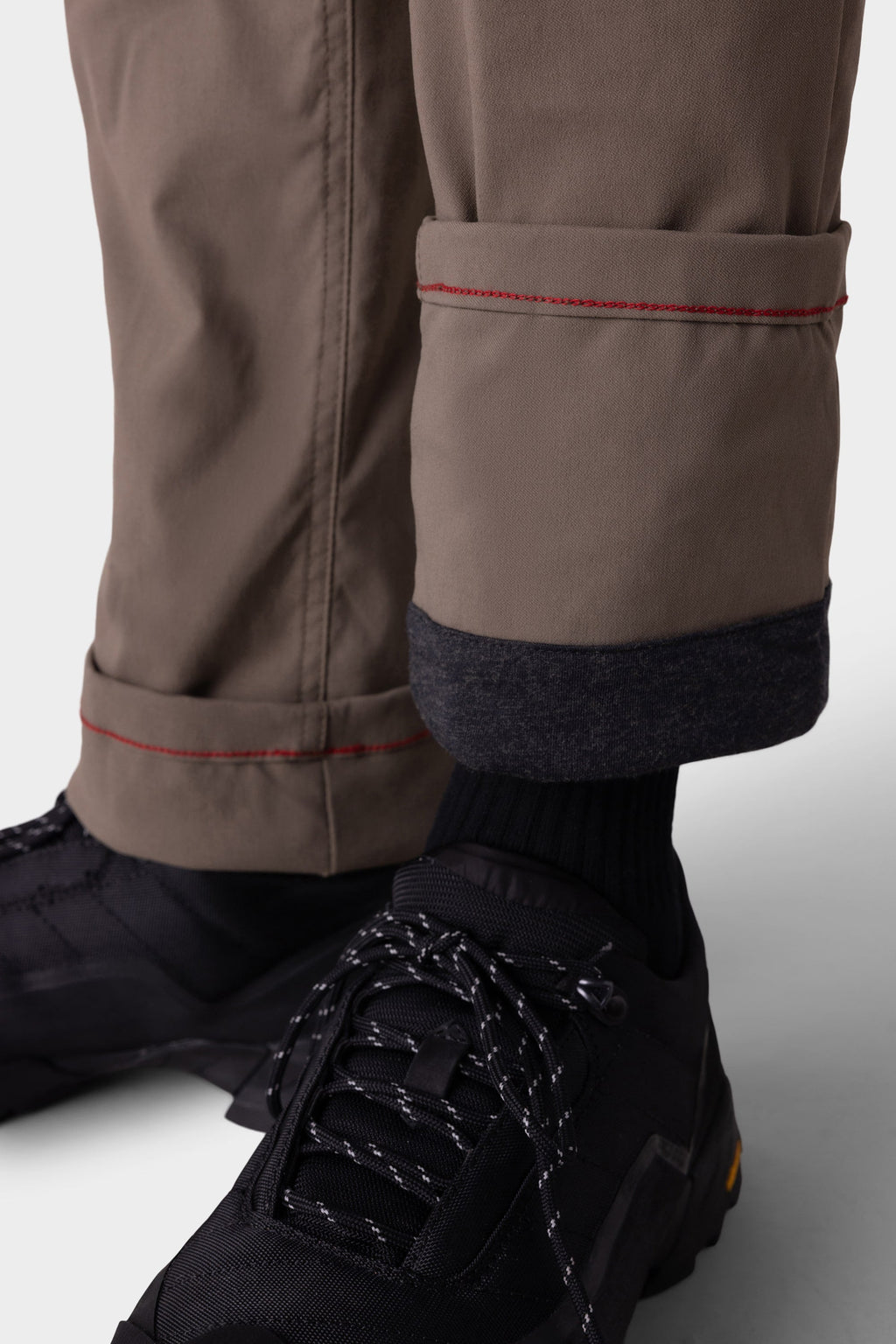 A close-up of a person wearing 686 Men's Everywhere® Merino-Lined Pant - Slim Fit in taupe with red stitching and rolled cuffs revealing a warm dark lining, paired with black socks and black hiking shoes with white speckled laces.