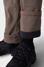 A close-up of a person wearing 686 Men's Everywhere® Merino-Lined Pant - Slim Fit in taupe with red stitching and rolled cuffs revealing a warm dark lining, paired with black socks and black hiking shoes with white speckled laces.