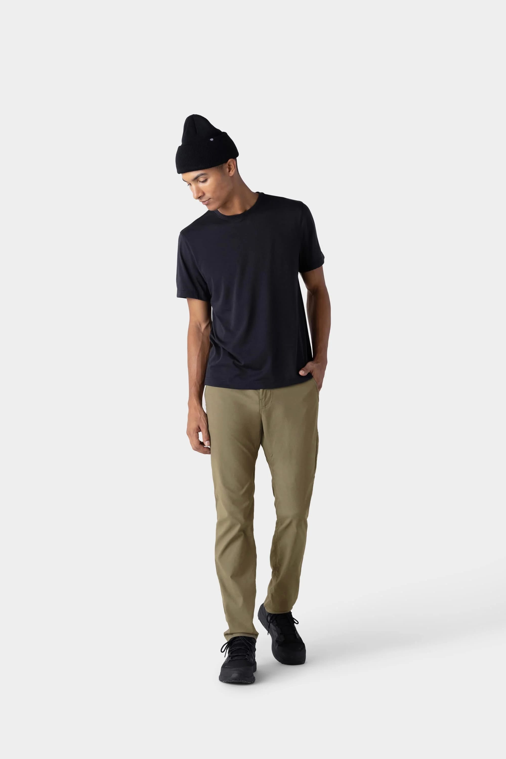 Wearing a black beanie, black t-shirt, black shoes, and 686 Men's Everywhere® Merino-Lined Pant - Slim Fit by 686 in khaki, a person stands against a plain white background, looking down with one hand in their pocket.