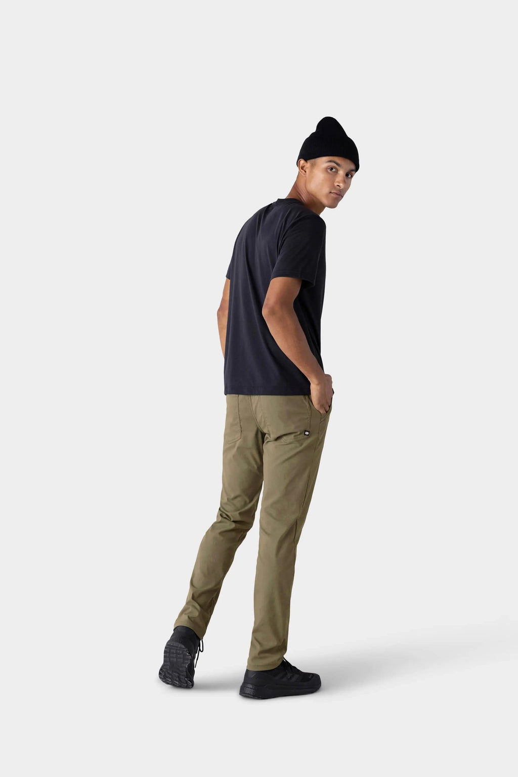 A man wearing a black beanie, black t-shirt, and 686 Men's Everywhere® Merino-Lined Pant - Slim Fit in olive stands against a plain light gray background, turning slightly to look over his shoulder.