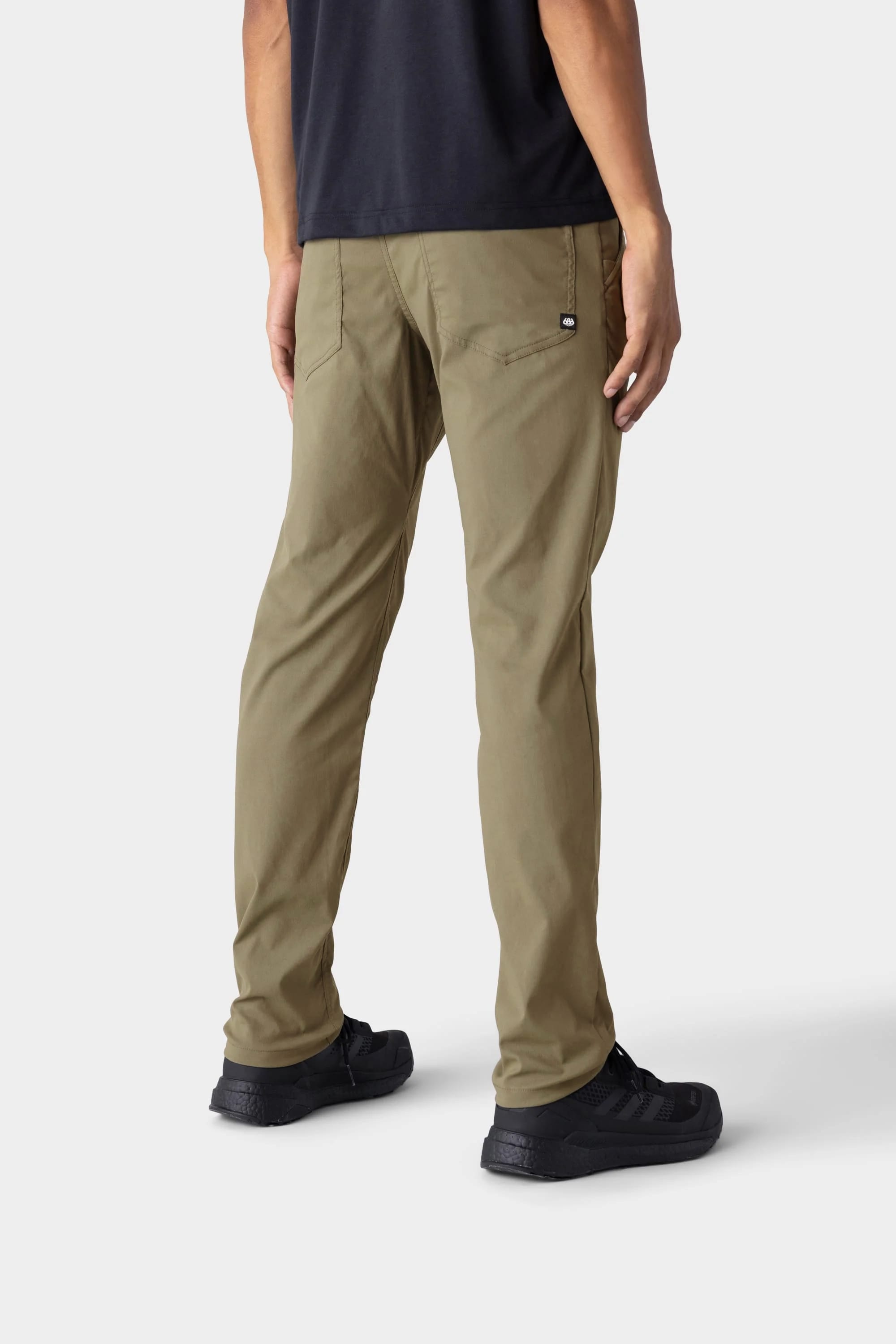 A person in 686 Men's Everywhere® Merino-Lined Pant - Slim Fit (olive), a black T-shirt, and black sneakers stands with their back to the camera, highlighting the pants against a plain light background.
