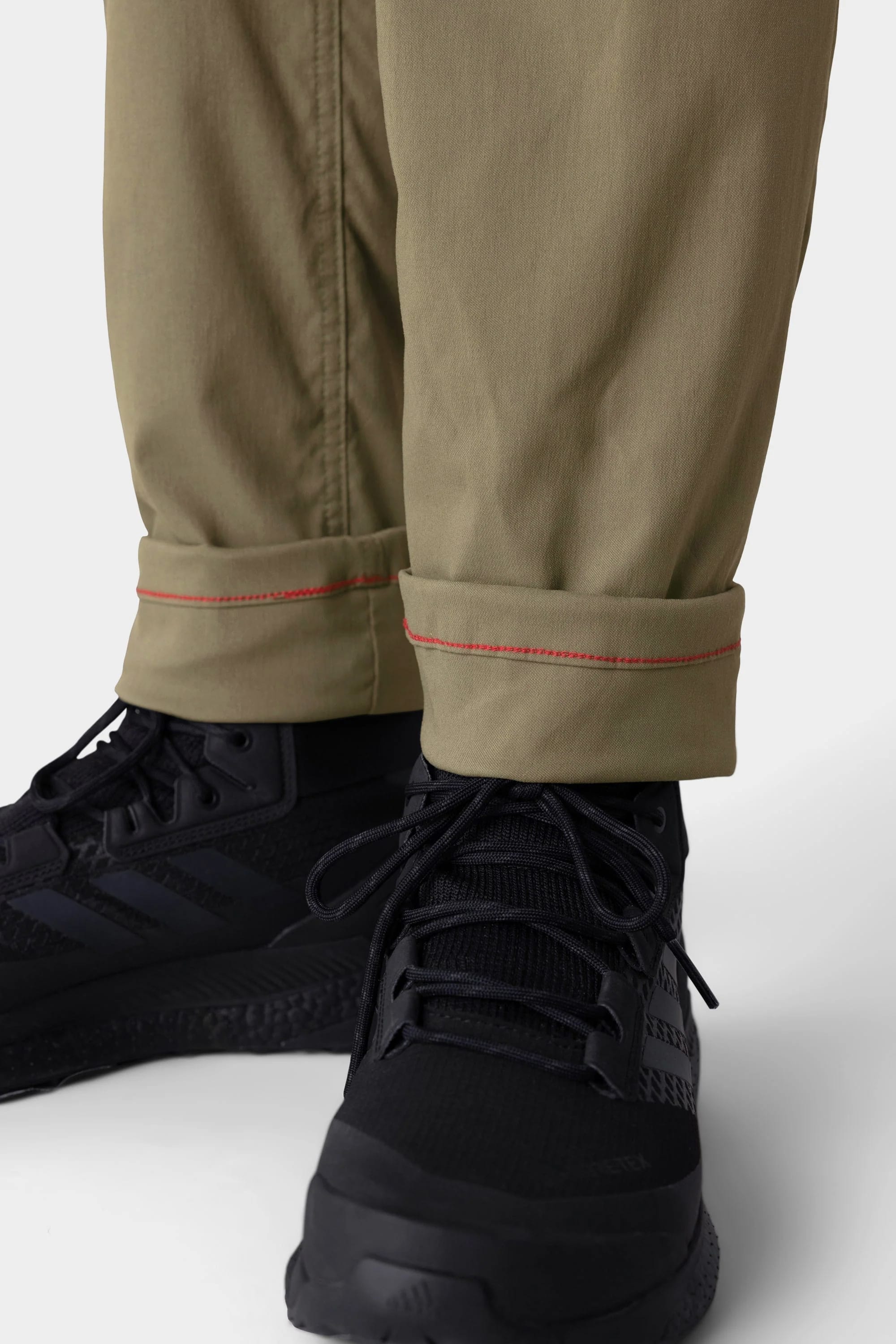 Close-up of a person wearing 686 Men's Everywhere® Merino-Lined Pant - Slim Fit with cuffed khaki hem featuring red stitching, paired with black athletic shoes—perfect for staying warm in cold weather. Focus is on the lower legs and feet.