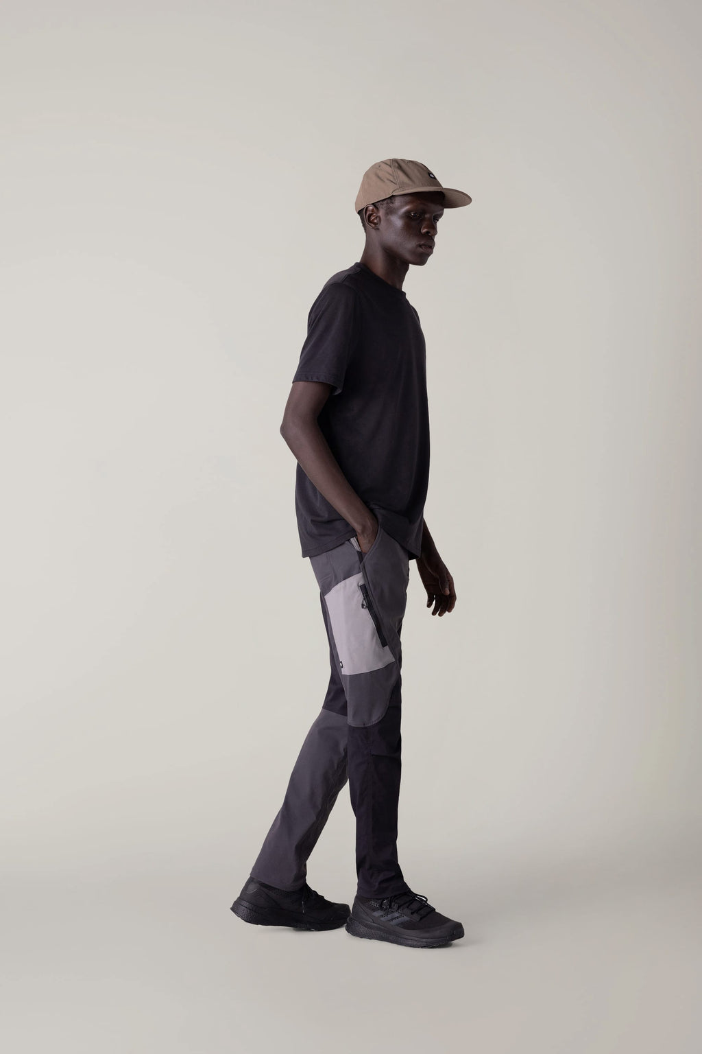 A man stands sideways against a plain light background, wearing a beige cap, black t-shirt, 686 Men's Anything Cargo Pant - Slim Fit by 686, and black shoes, his left hand in his pocket—ready for any adventure.
