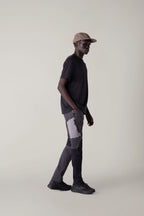 A man stands sideways against a plain light background, wearing a beige cap, black t-shirt, 686 Men's Anything Cargo Pant - Slim Fit by 686, and black shoes, his left hand in his pocket—ready for any adventure.
