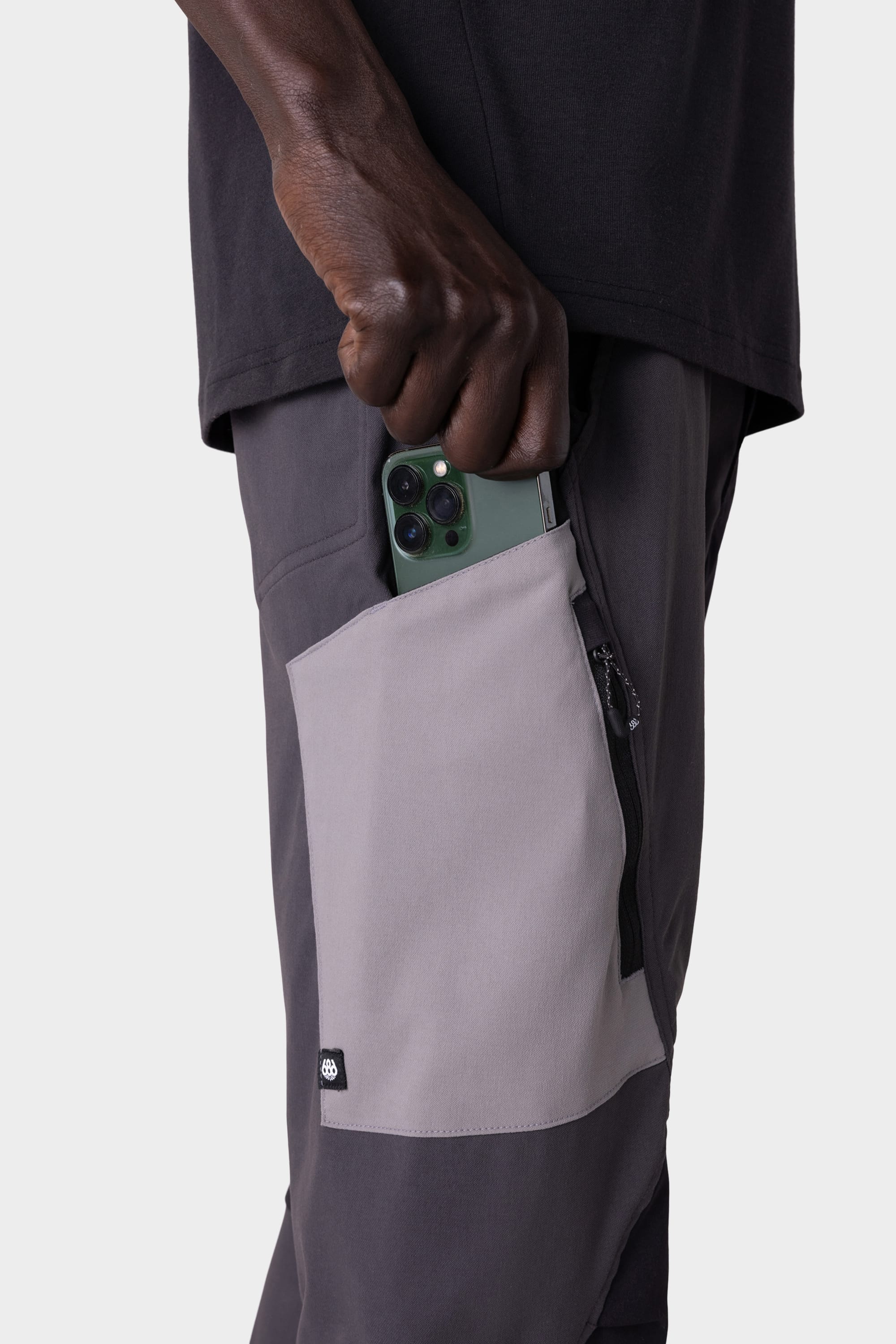 Wearing the 686 Men's Anything Cargo Pant - Slim Fit, a person in a black shirt slips a green smartphone into a light gray zippered side pocket on these 13-pocket pants, ready for any adventure.