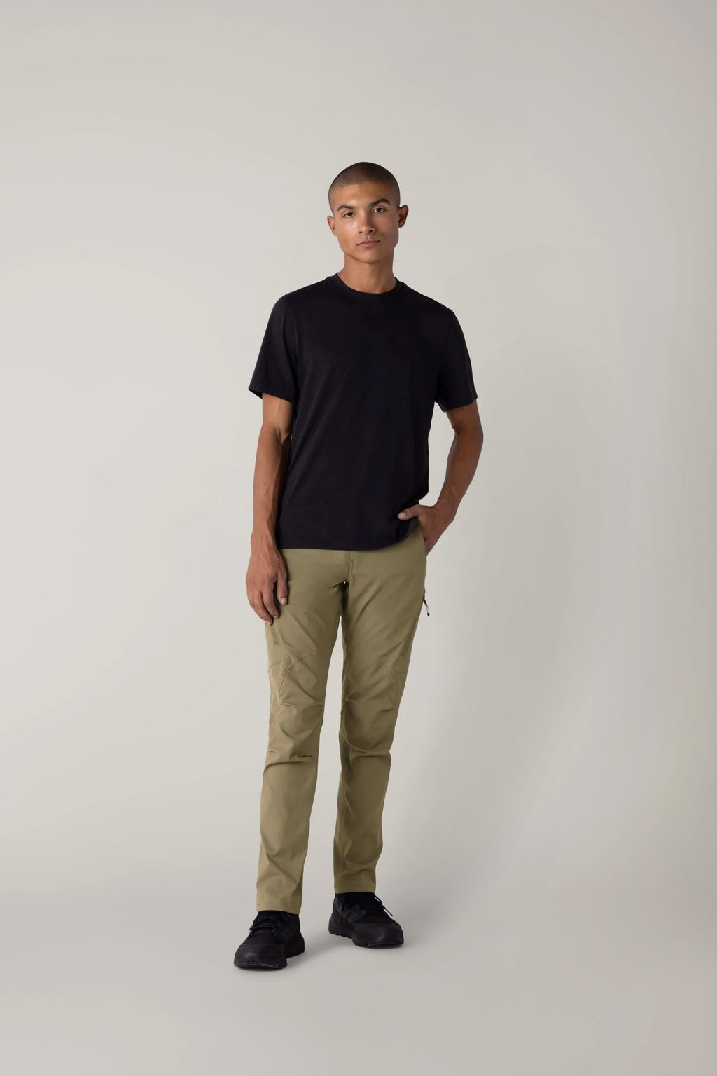 Against a plain light background, a person wears a black T-shirt, black shoes, and the 686 Men's Anything Cargo Pant - Slim Fit by 686, hand in pocket, projecting an understated, adventure-ready vibe while looking at the camera with a neutral expression.