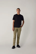 Against a plain light background, a person wears a black T-shirt, black shoes, and the 686 Men's Anything Cargo Pant - Slim Fit by 686, hand in pocket, projecting an understated, adventure-ready vibe while looking at the camera with a neutral expression.