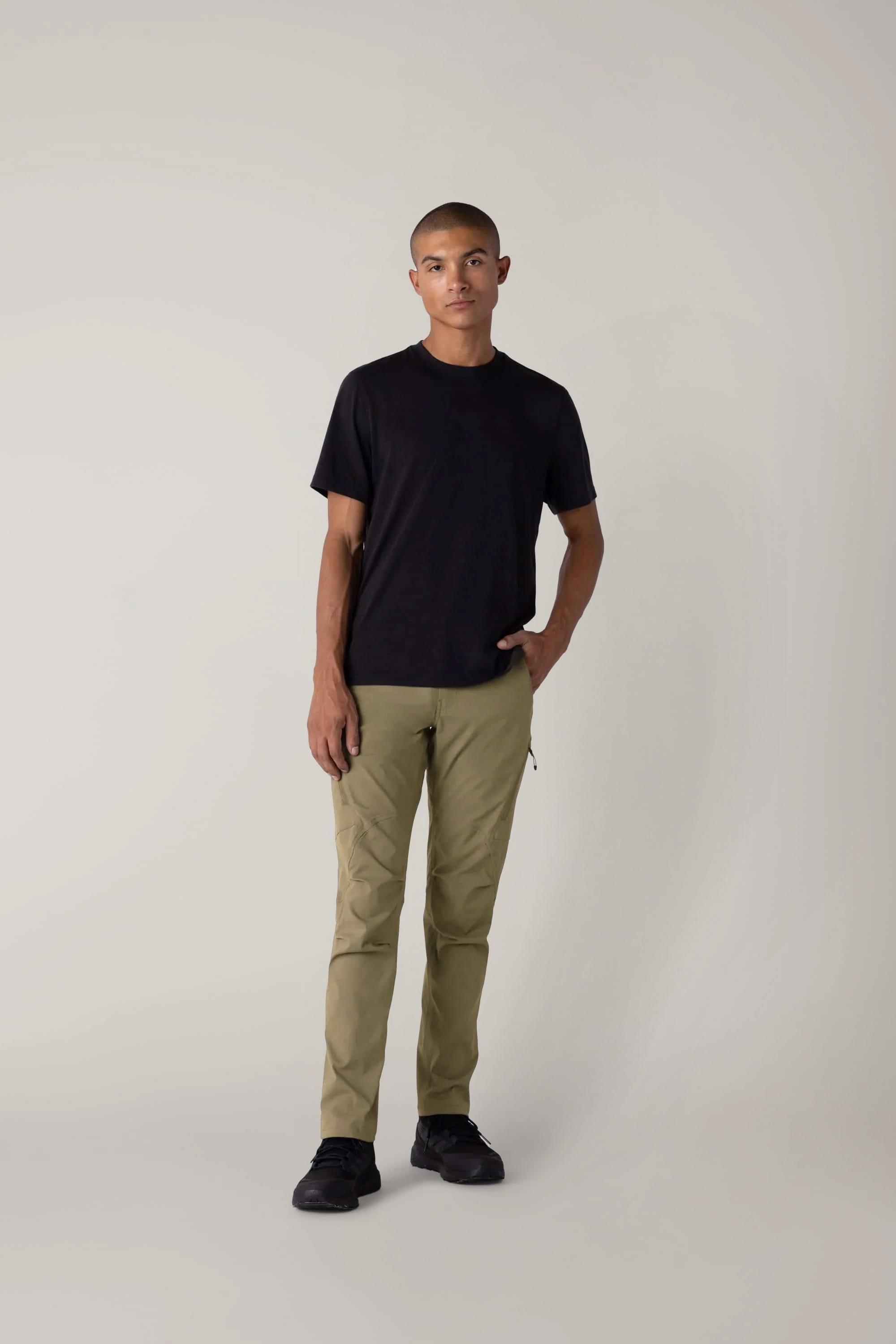 Against a plain light background, a person wears a black T-shirt, black shoes, and the 686 Men's Anything Cargo Pant - Slim Fit by 686, hand in pocket, projecting an understated, adventure-ready vibe while looking at the camera with a neutral expression.