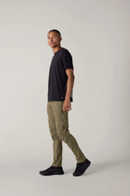 A man with short hair wears a black t-shirt, 686 Men's Anything Cargo Pant - Slim Fit in olive green by 686, and black shoes, standing in profile against a plain light gray background, ready for any adventure.