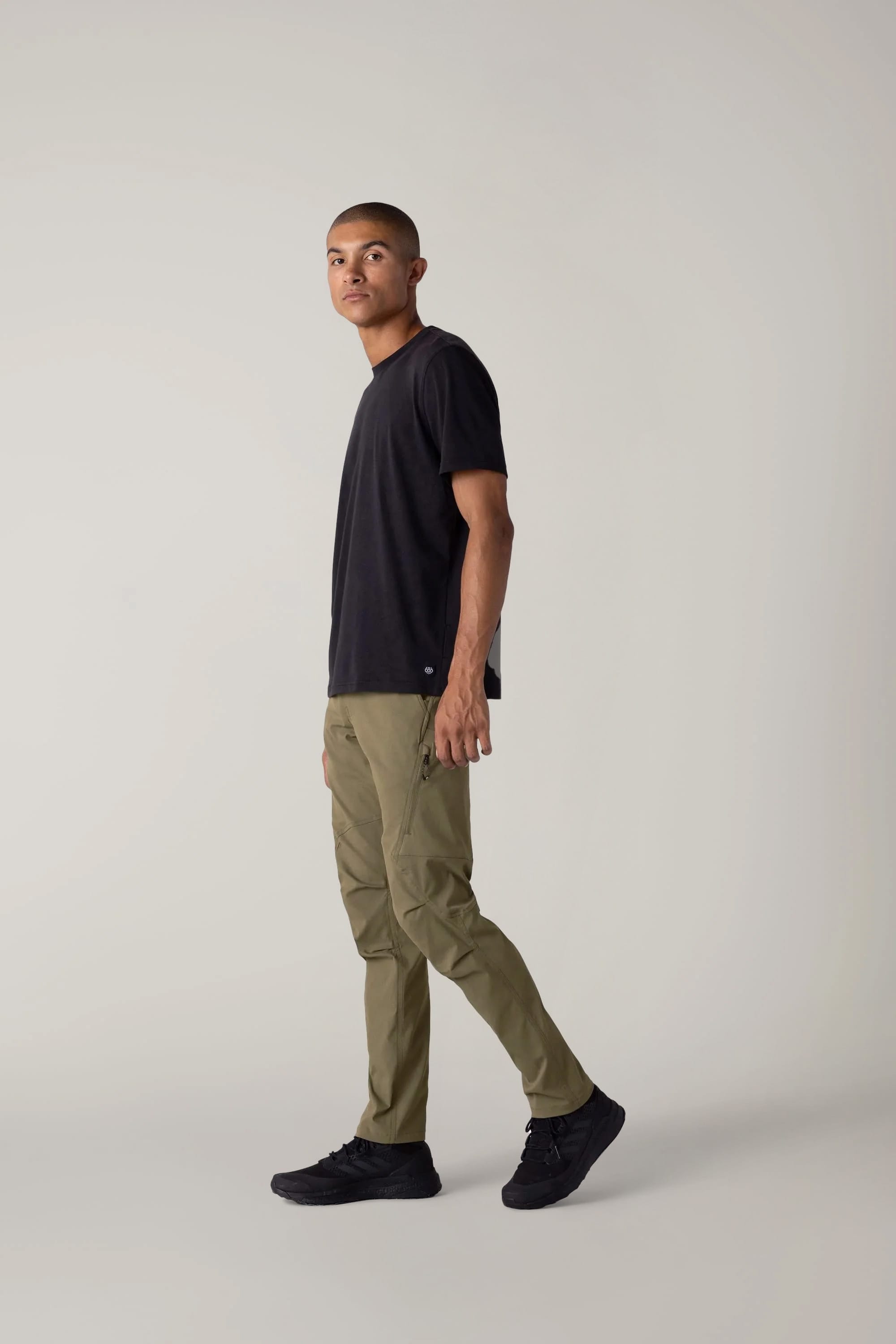 A man with short hair wears a black t-shirt, 686 Men's Anything Cargo Pant - Slim Fit in olive green by 686, and black shoes, standing in profile against a plain light gray background, ready for any adventure.
