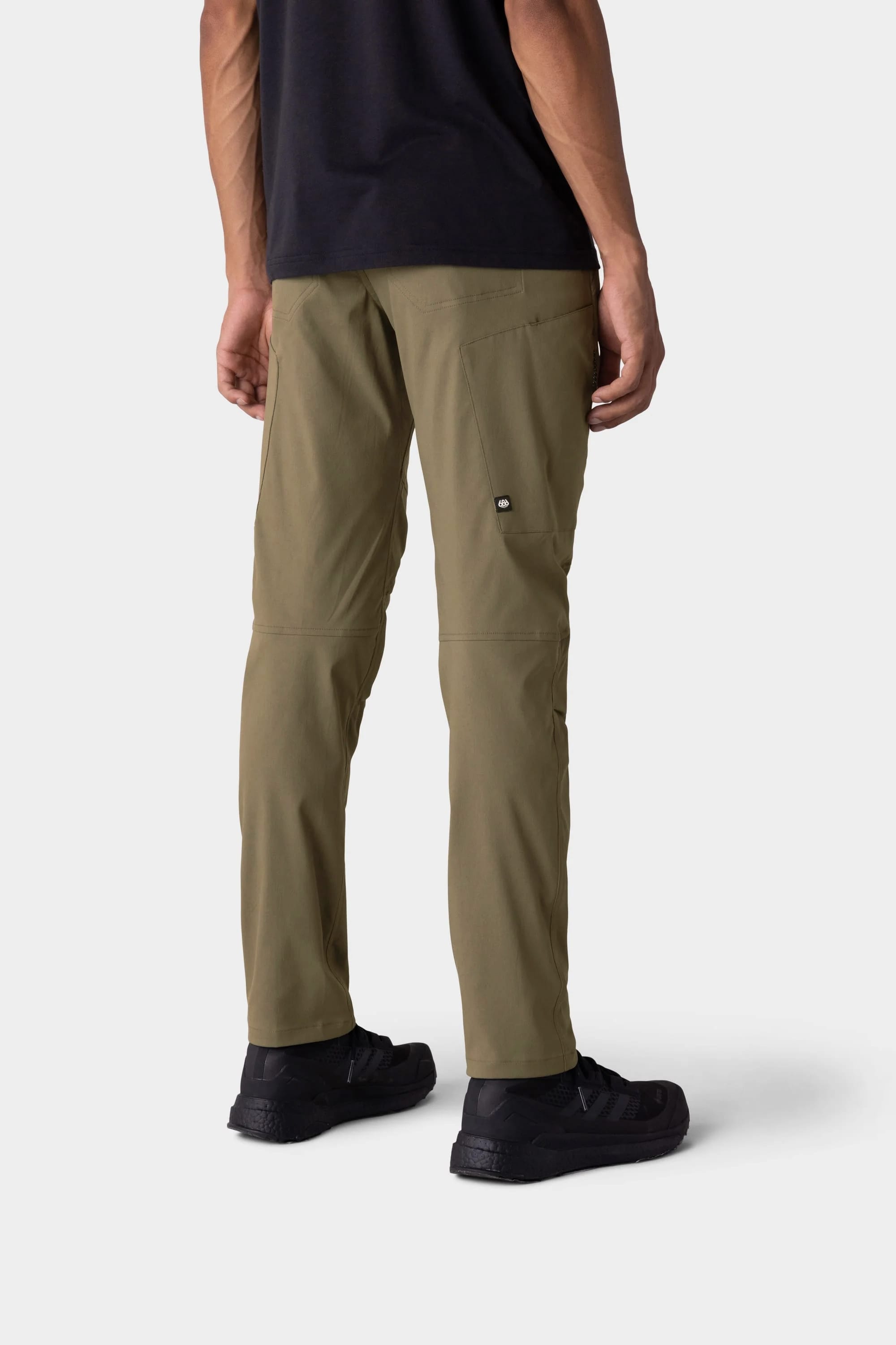 A person stands with their back to the camera against a light gray background, wearing 686 Men's Anything Cargo Pant - Slim Fit in olive green, paired with a black shirt and sneakers—perfectly equipped for any adventure.