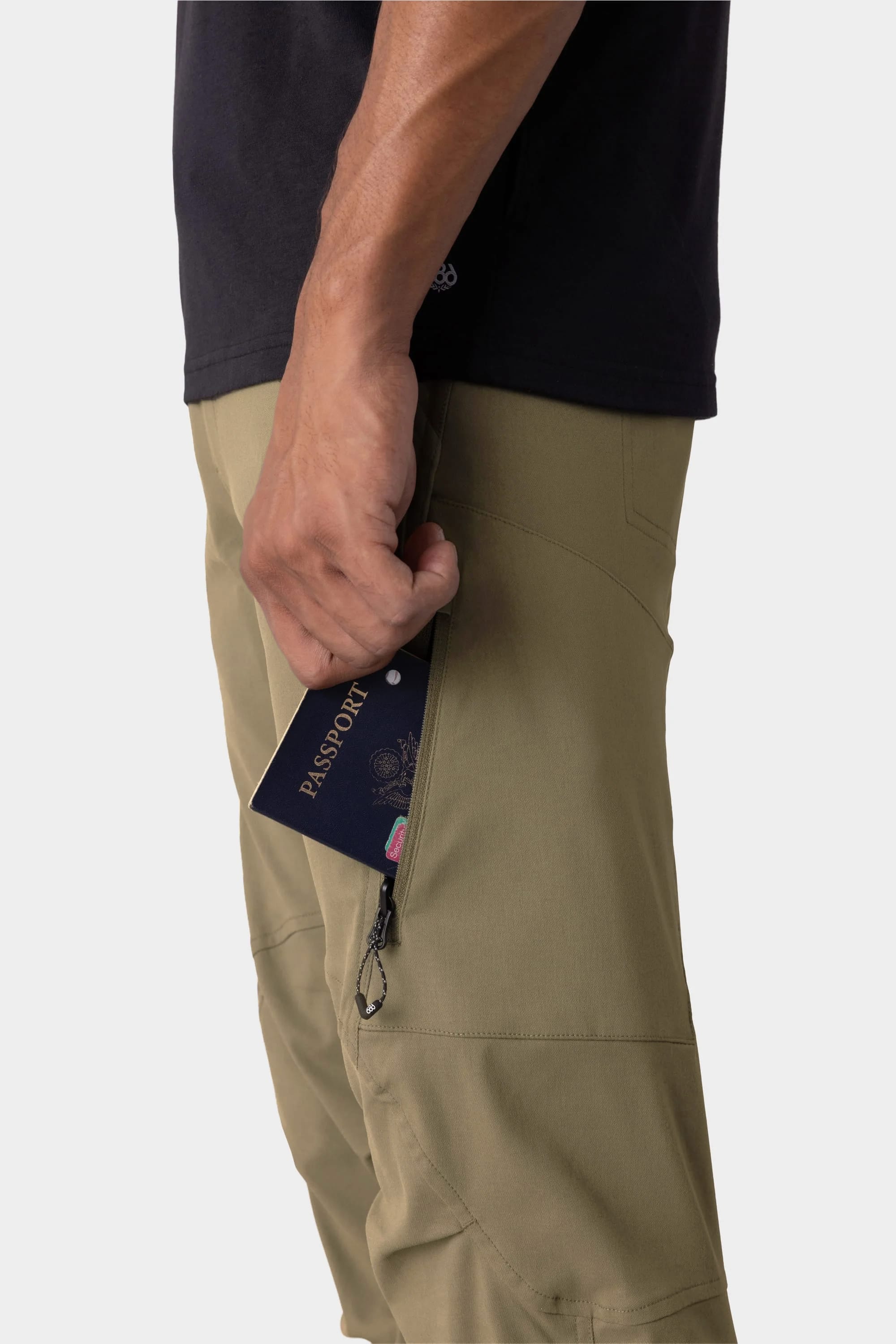 A person slips a passport into the zippered side pocket of 686 Men's Anything Cargo Pant - Slim Fit. Only their arm, hand, and part of their torso and legs in a black shirt are visible. Brand: 686.