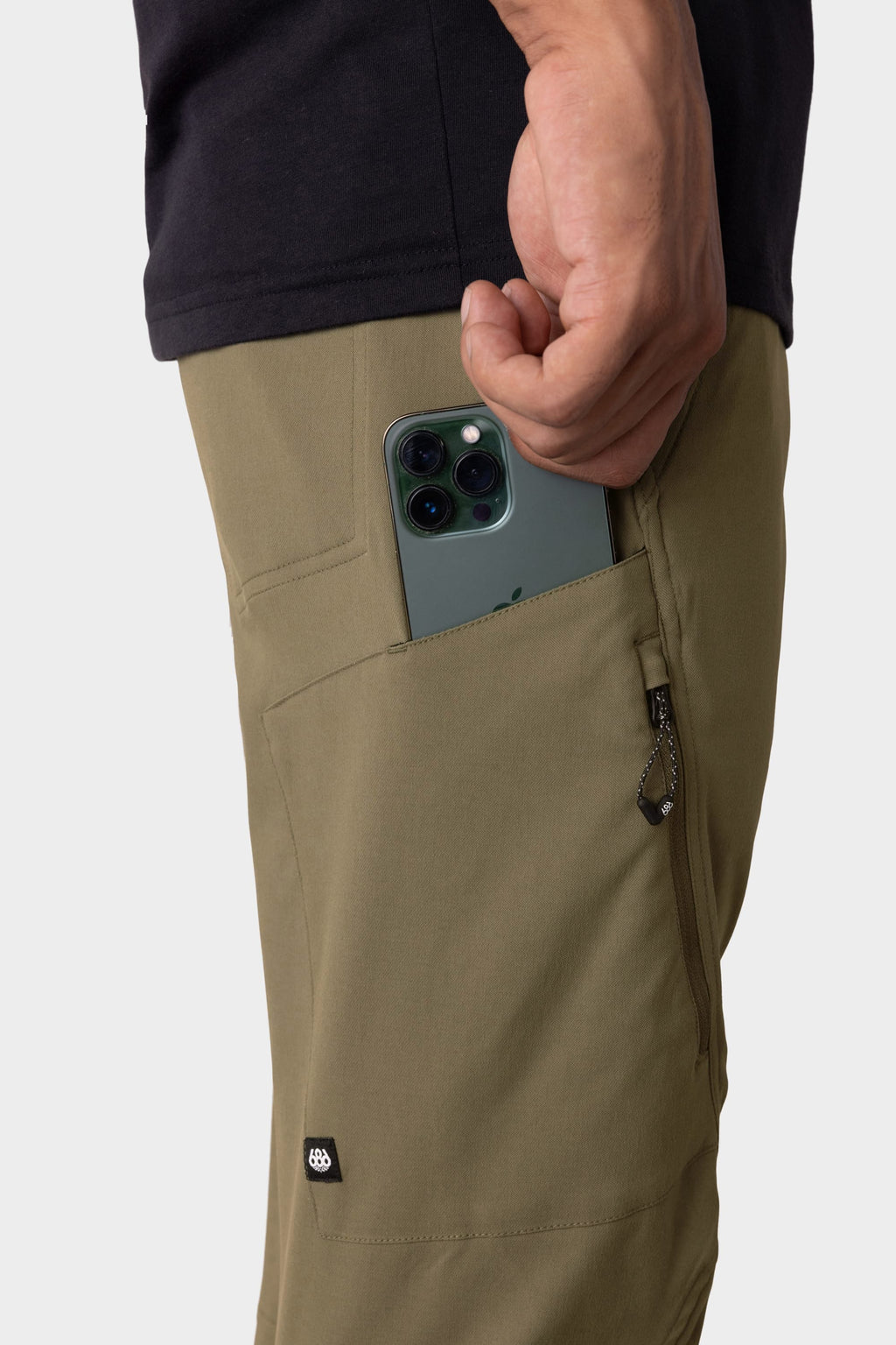 Wearing 686 Men's Anything Cargo Pant - Slim Fit and a black shirt, a person slips a green triple-camera smartphone into a zippered side pocket, ready for any adventure.