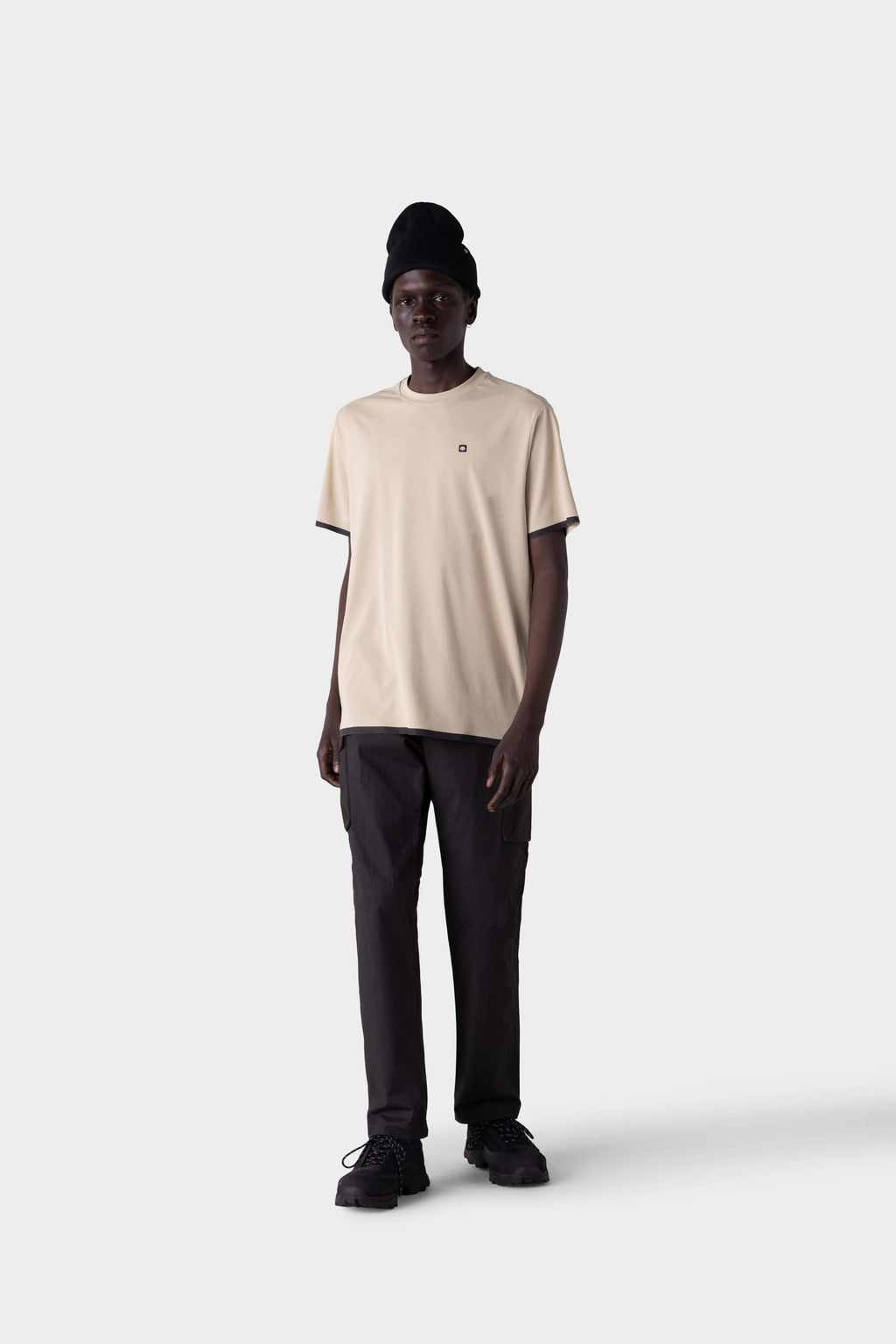A person stands facing forward against a light gray background, wearing the 686 Men's Let's Go Tech Tee in beige with black pants, shoes, and beanie—a perfect look from 686 for spring summer adventure.