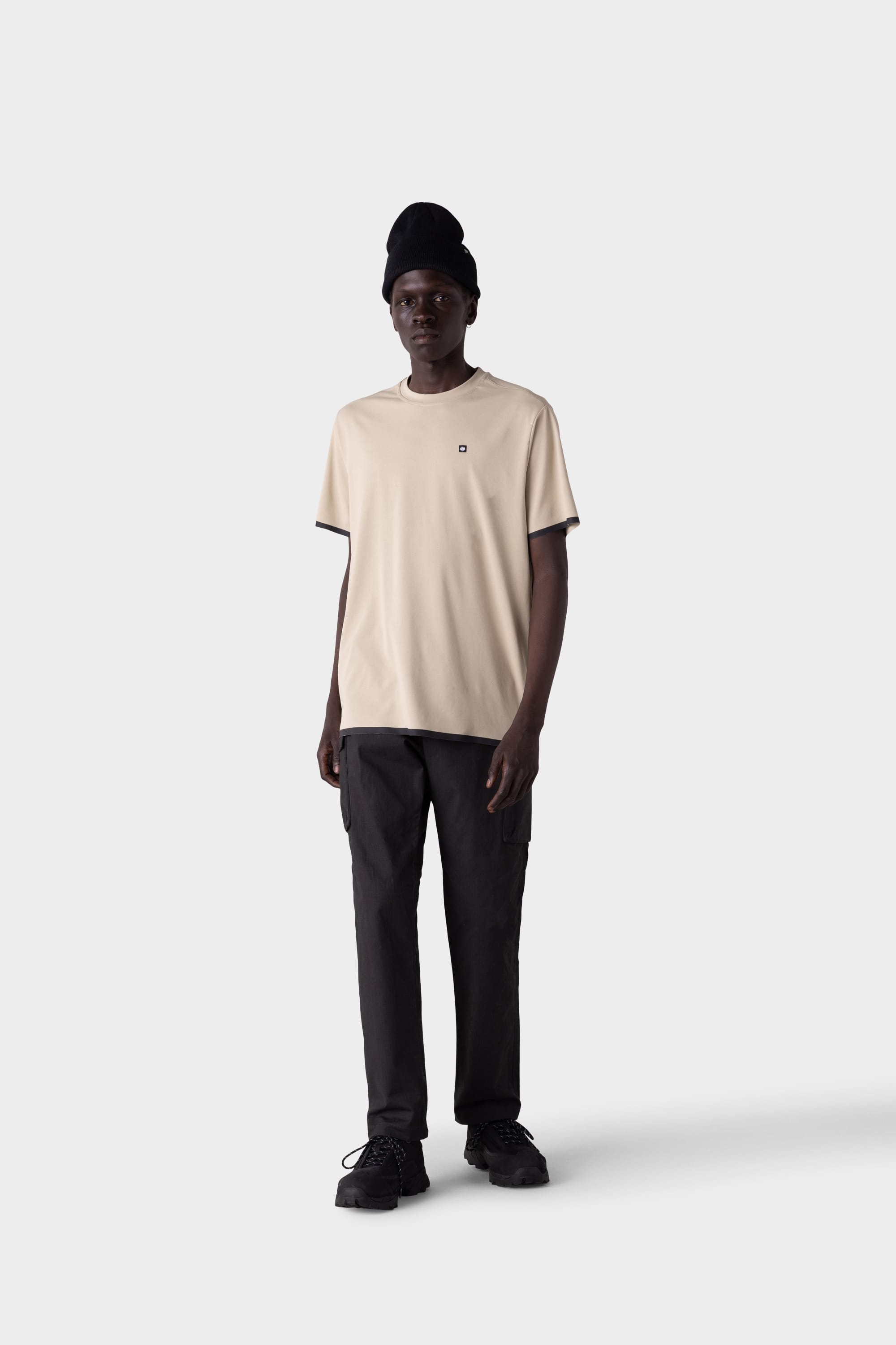 A person stands facing forward against a light gray background, wearing the 686 Men's Let's Go Tech Tee in beige with black pants, shoes, and beanie—a perfect look from 686 for spring summer adventure.