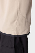 Close-up of a person in a beige textured jacket with a hidden side pocket and black trim, layered over the 686 Men's Let's Go Tech Tee by 686, paired with black cargo pants. The background is plain and light-colored.