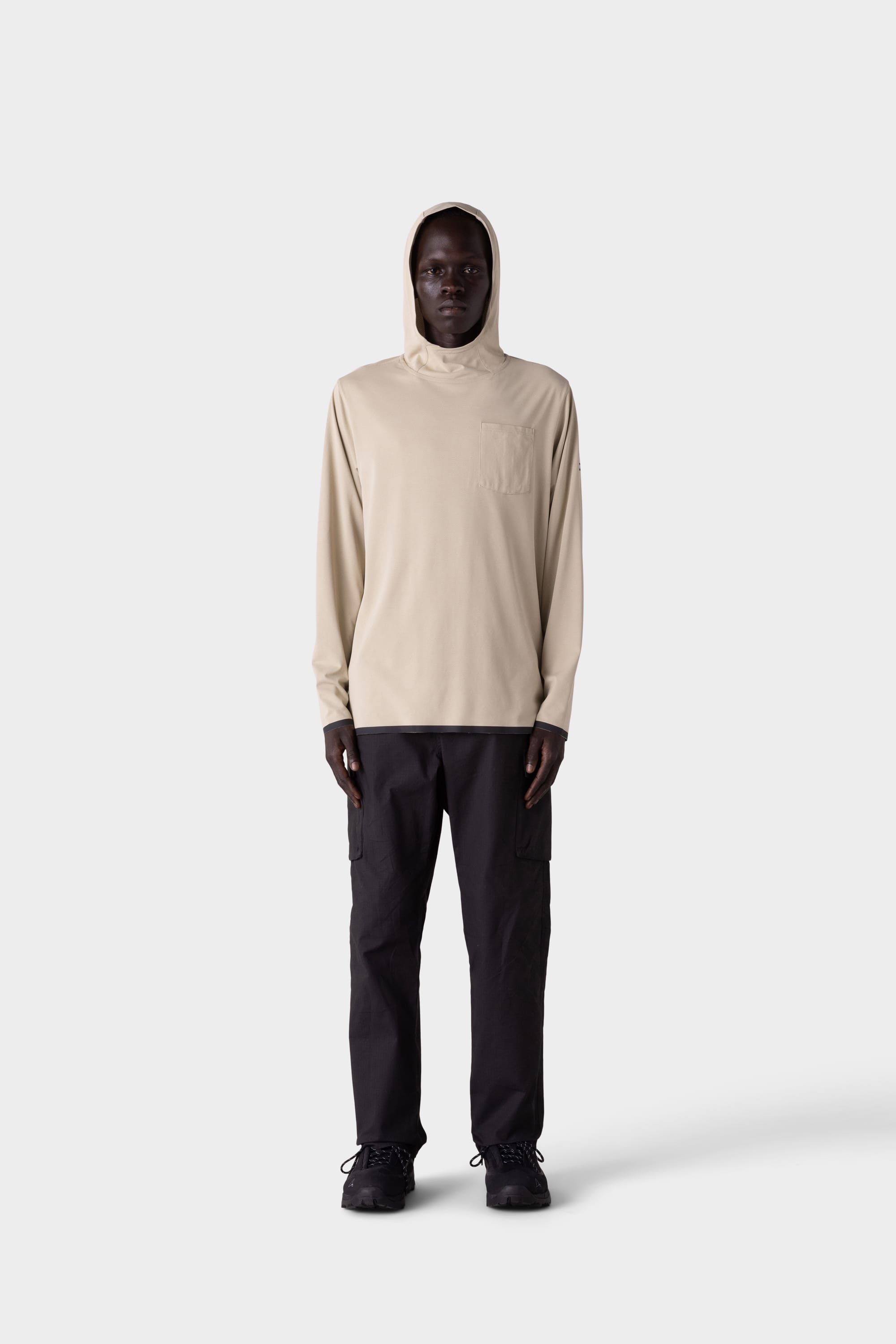 A person stands facing forward wearing the 686 Mens Lets Go Tech Hoody in light beige, paired with black cargo pants and black shoes, against a plain light gray background.