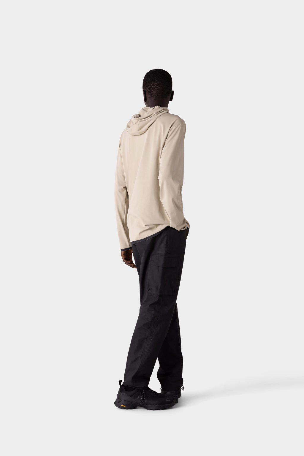 A person stands facing away from the camera in a light beige 686 Mens Lets Go Tech Hoody—lightweight and breathable for sun protection—with loose black pants and black shoes, against a plain light gray background.