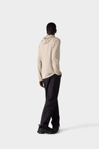 A person stands facing away from the camera in a light beige 686 Mens Lets Go Tech Hoody—lightweight and breathable for sun protection—with loose black pants and black shoes, against a plain light gray background.
