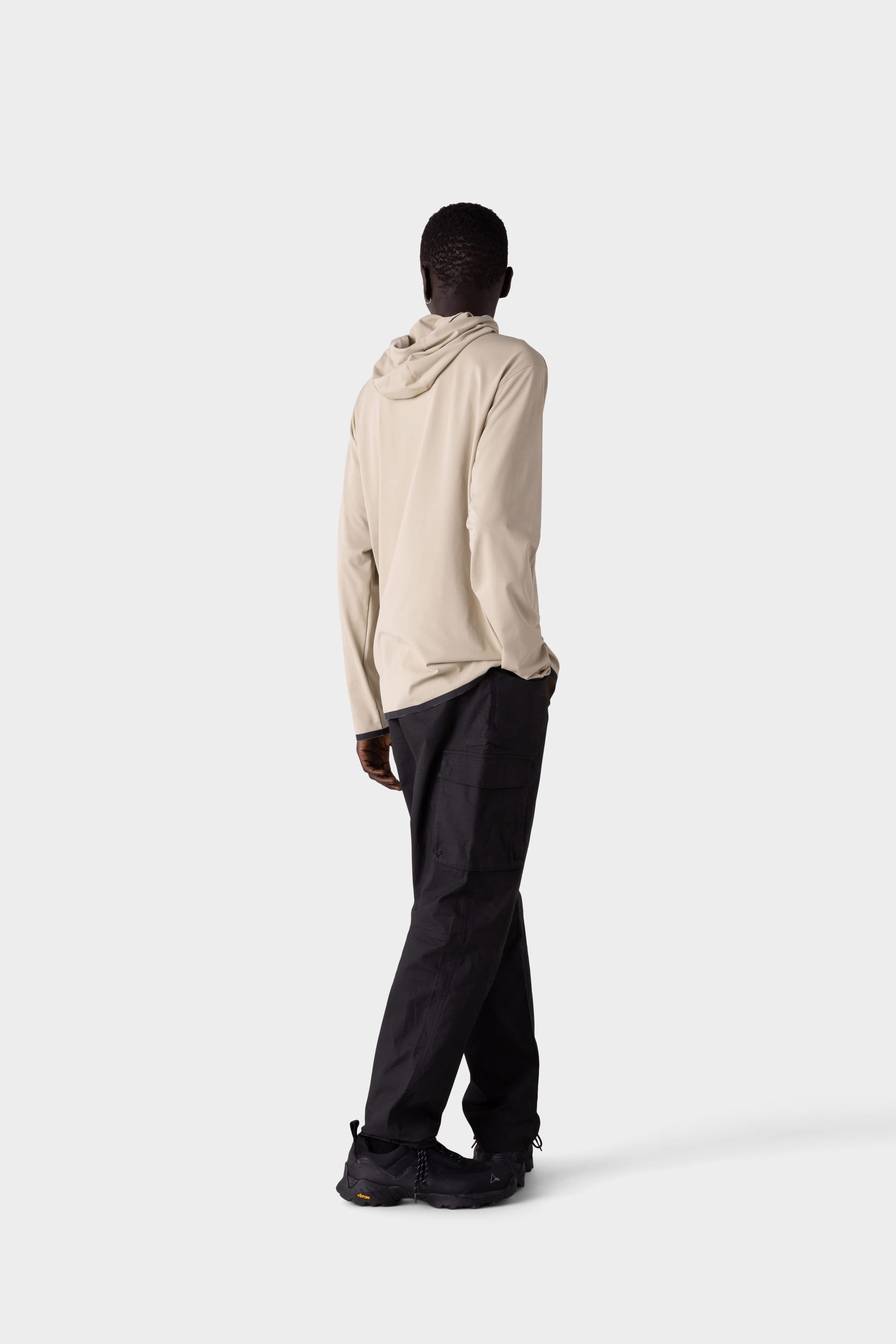 A person stands facing away from the camera in a light beige 686 Mens Lets Go Tech Hoody—lightweight and breathable for sun protection—with loose black pants and black shoes, against a plain light gray background.