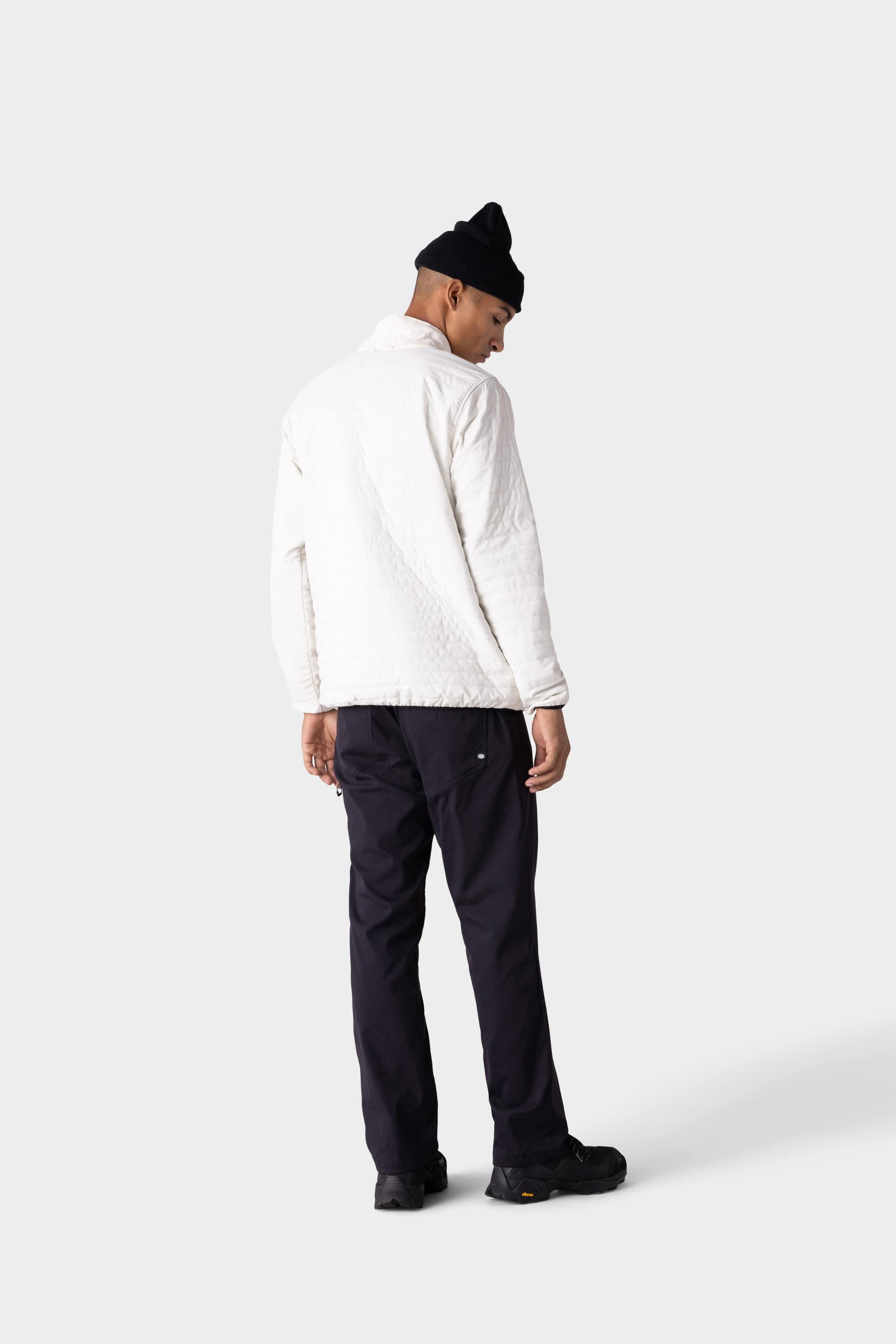 A man faces away, wearing black 686 Men's Everywhere® Merino-Lined Pant - Relaxed Fit, a white quilted coat, black shoes, and a black beanie against a plain light gray background.