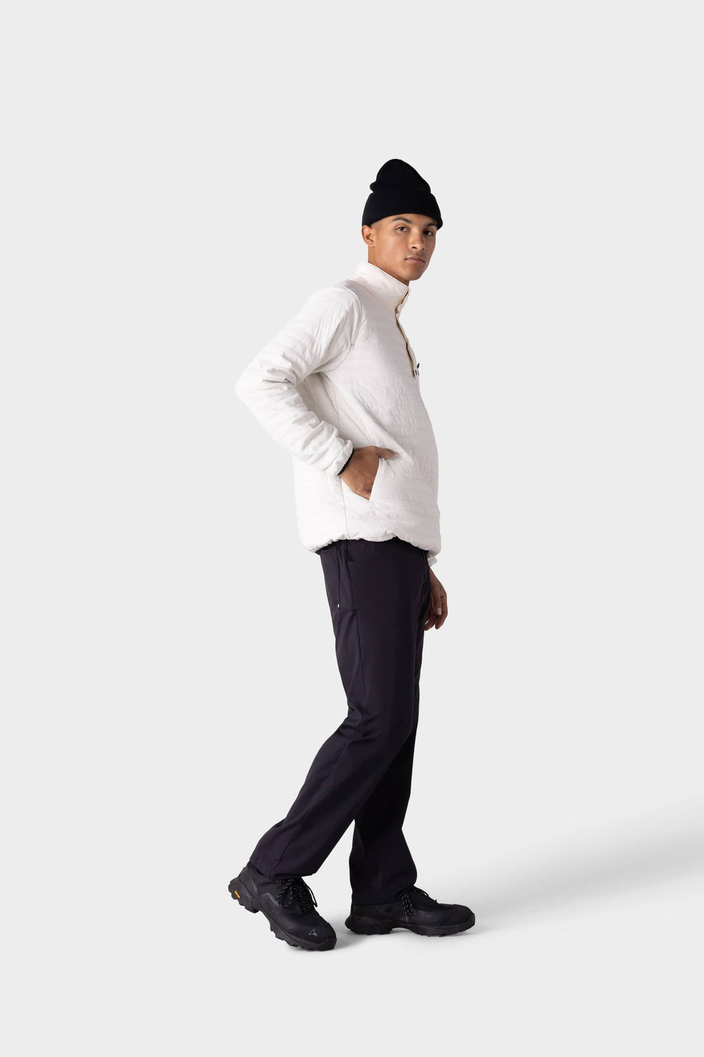 A person wearing a white long-sleeve shirt, black shoes, a black beanie, stylish insulated outerwear, and the 686 Men's Everywhere® Merino-Lined Pant - Relaxed Fit by 686 poses against a plain light gray background.