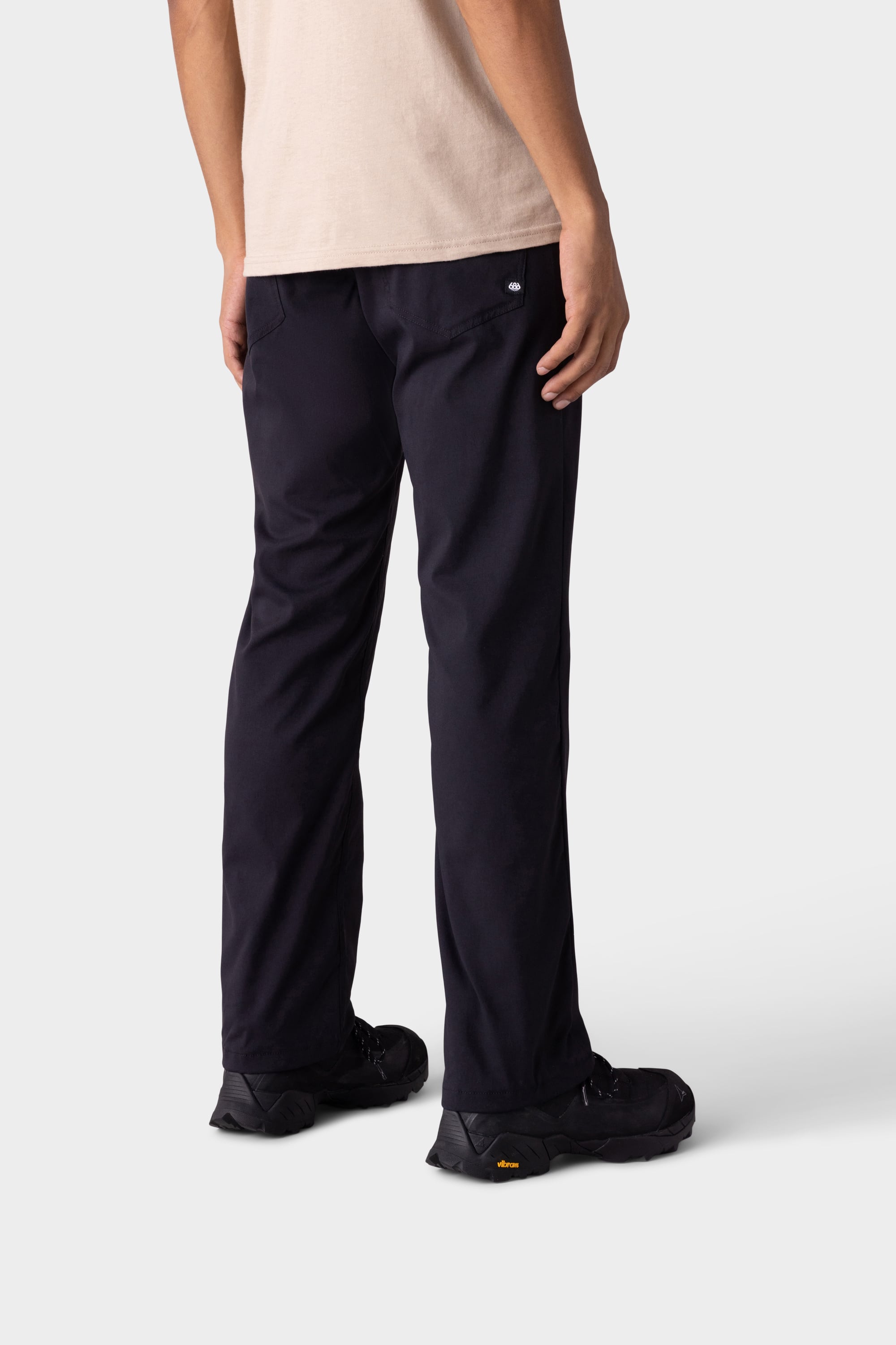 A person stands facing away from the camera against a plain light background, wearing black 686 Men's Everywhere® Merino-Lined Pant - Relaxed Fit, black shoes, a warm winter jacket, and a light beige short-sleeve shirt.