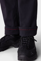 Close-up of a person wearing the 686 Men's Everywhere® Merino-Lined Pant - Relaxed Fit, styled with a warm winter jacket and black athletic shoes. The background is plain and light-colored.