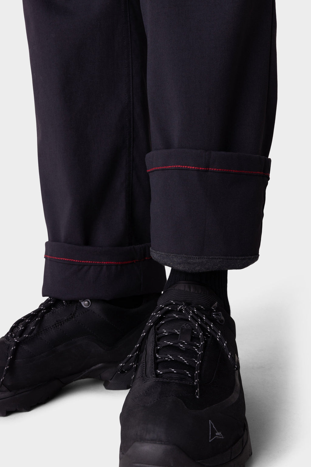 A close-up shows a person wearing 686 Men's Everywhere® Merino-Lined Pant - Relaxed Fit, featuring black fabric with red-stitched cuffs, paired with black athletic shoes and rolled up to reveal dark socks—completing a stylish insulated look.