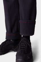 A close-up shows a person wearing 686 Men's Everywhere® Merino-Lined Pant - Relaxed Fit, featuring black fabric with red-stitched cuffs, paired with black athletic shoes and rolled up to reveal dark socks—completing a stylish insulated look.