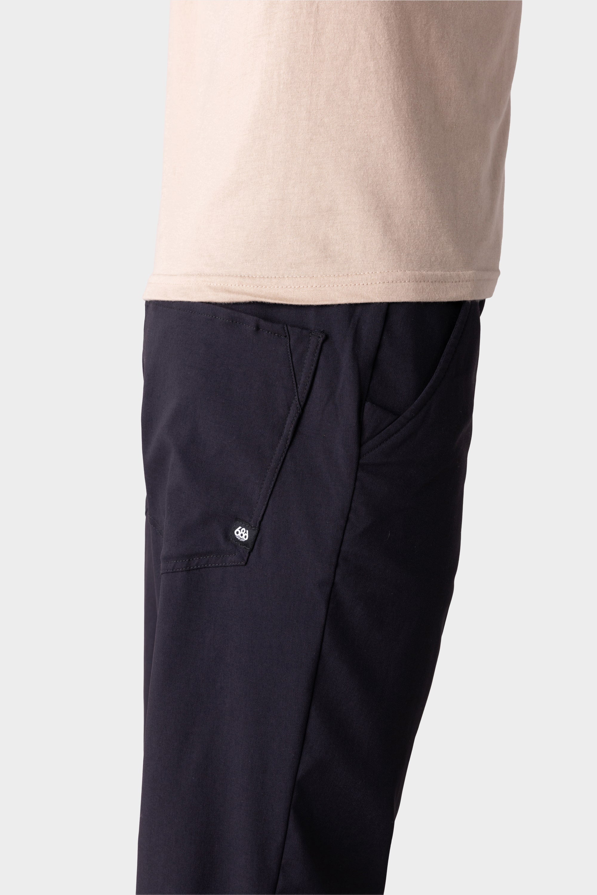 A person in 686 Men's Everywhere® Merino-Lined Pant - Relaxed Fit, shown from the waist down and side view, pairs dark pants with a beige t-shirt and insulated outerwear, hand in pocket.