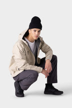 A person wearing a beige insulated jacket, white shirt, black beanie, 686 Men's Everywhere® Merino-Lined Pant - Relaxed Fit in charcoal gray, and black sneakers kneels on one knee against a plain light gray background.