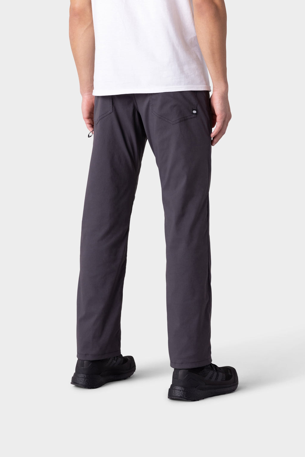 A person is shown from the back wearing insulated outerwear, a white t-shirt, and 686 Men's Everywhere® Merino-Lined Pant - Relaxed Fit in dark gray, paired with black sneakers against a plain light gray background.